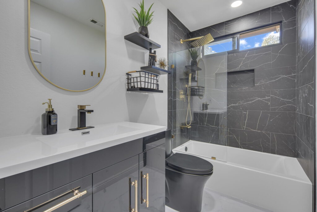 Modern bathroom with dark gray vanity, gold hardware, and a round gold-framed mirror above a white countertop.