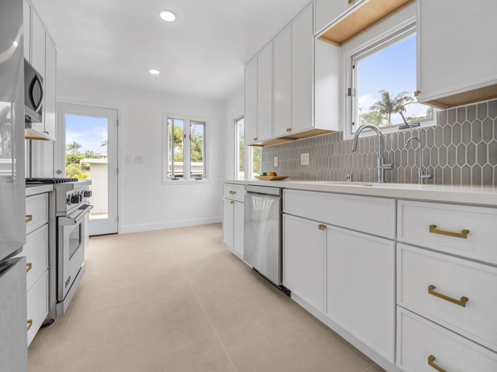 Bright modern kitchen with white cabinets, stainless steel appliances, and a gray hex tile backsplash bathed in natural light from large windows.
