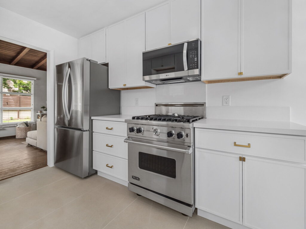 Modern kitchen with white cabinets and stainless steel appliances including a side-by-side refrigerator, microwave, and gas range under a range hood in bright space.